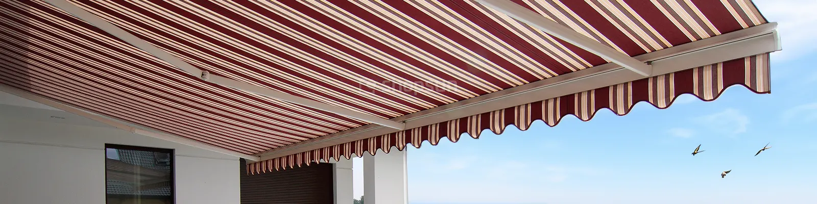 Große Terrassenmarkise mit drei Armen und bordeaux-beige gestreiftem Stoff vor einem blauen Himmel mit drei Schwalben im Hintergrund.